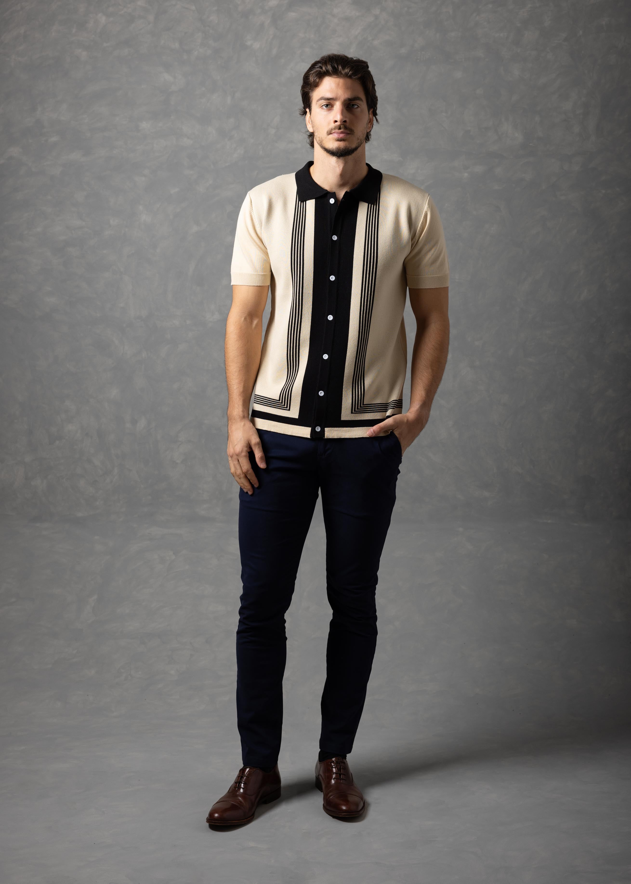 Man wearing a patterned shirt over a black dress shirt with dark jeans and brown shoes on a gray background