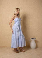 Woman in a light blue patterned dress standing next to a beige vase against a beige wall.